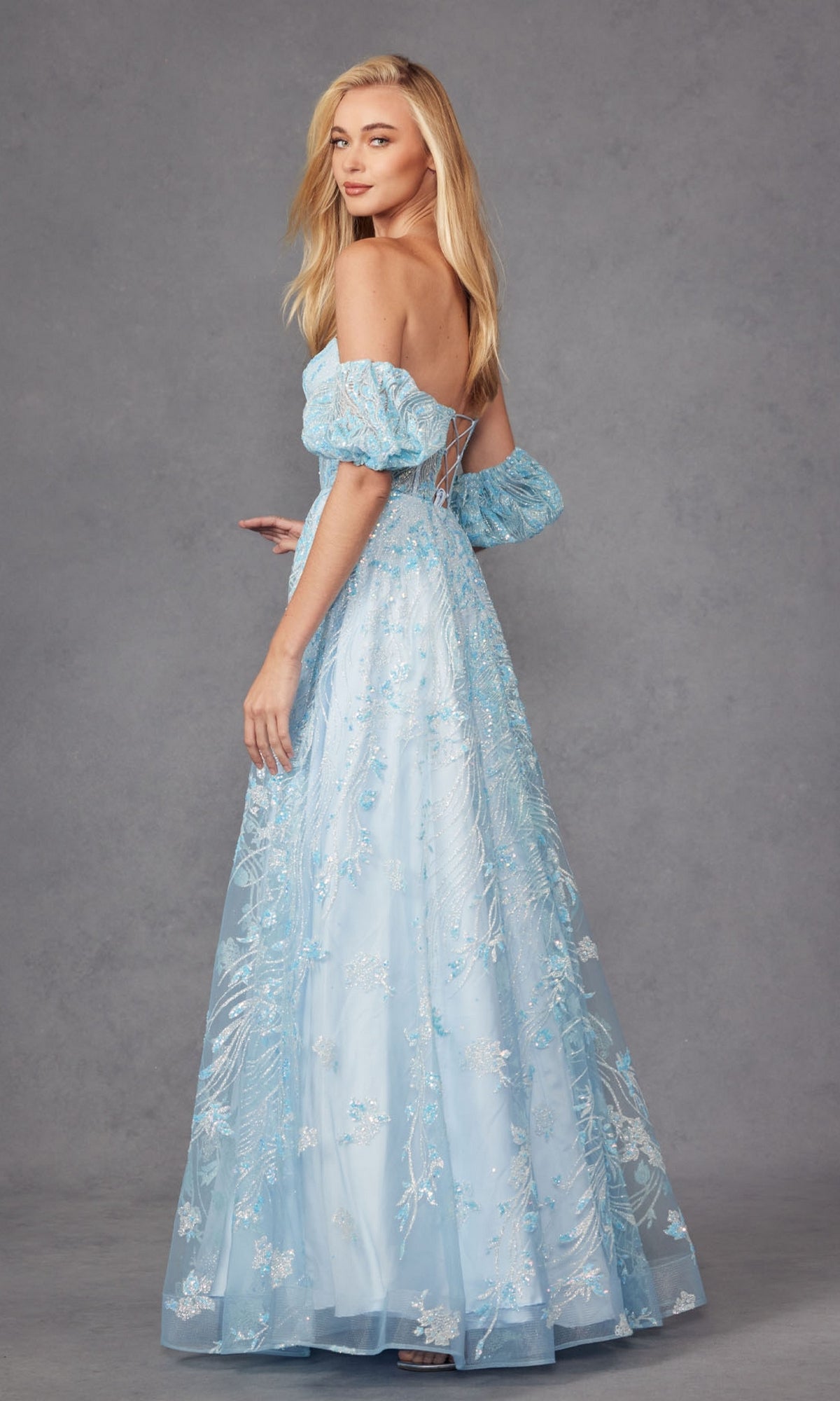 A woman poses against a plain gray background, wearing the Puff-Sleeve Strapless Long Glitter Prom Dress 2404 in light blue, looking over her shoulder.
